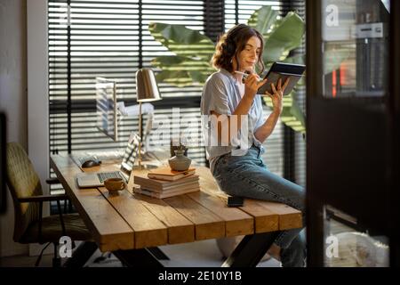 Young woman works on a touchpad, feeling comfortable at cozy home office with a green plant on the background Stock Photo