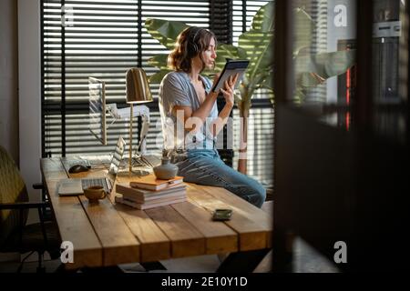 Young woman works on a touchpad, feeling comfortable at cozy home office with a green plant on the background Stock Photo