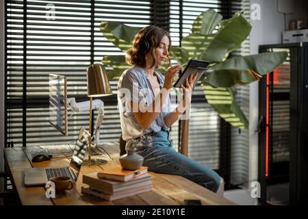 Young woman works on a touchpad, feeling comfortable at cozy home office with a green plant on the background Stock Photo