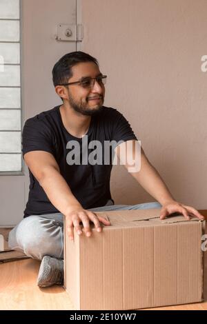 latin man opening a box in his new apartment by a window wearing black t-shirt and sportswear Stock Photo