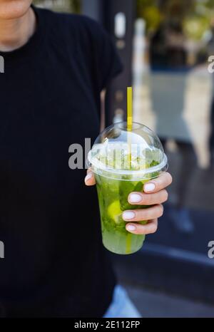 Female hand holding plastic cup with a mojito in a city cafe. Fresh cocktail drink with ice fruit and herb decoration. Selective focus, blurred backgr Stock Photo