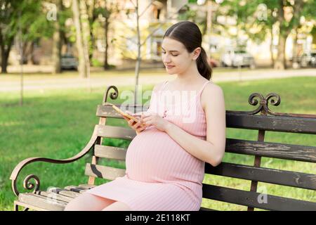 pregnant woman sits on a park bench and texting on phone. Stock Photo