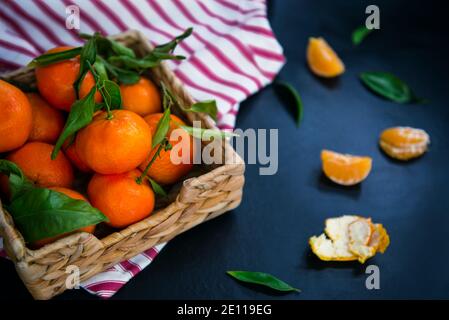 Fresh ripe mandarins, clementine, tangerine with green leaves in the straw bowl on a striped napkin and dark blue stone background. Local citrus fruit Stock Photo