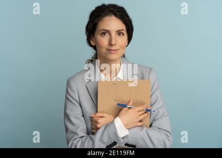 Business woman wear grey blazer presses clipboard to itself and looking at camera, studio blue wall Stock Photo