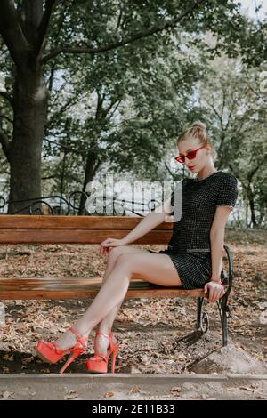 Pretty blonde girl sits on a bench in a park; she is wearing a stylish short dark dress, red sunglasses and high heels Stock Photo