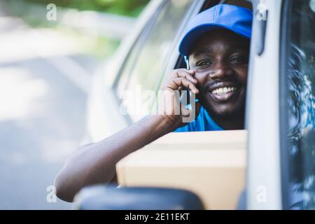 African postal delivery courier man using smart phone and delivering package in car Stock Photo