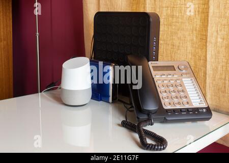 home office set up with a voice activated, virtual smart assistant, a landline telephone, and two routers on a white desk with  a burgundy and yellow Stock Photo