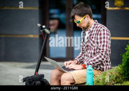 Young caucasian man information technology specialist works laptop outdoor, sitting on bench near house, alternative transport electric scooter stands Stock Photo