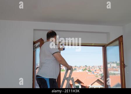 Man on a ladder painting a wall above a window in white color with a roller. Window is open and and red roofs could be seen. Stock Photo