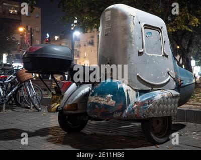 OLD CLASSIC VESPA SCOOTER WITH COVERED SIDECAE SEAT PARKING IN URBAN PARKING LOCATION AT NIGHT. THE SIDE CAR HAS WINDOW AND IT LOOKS LIKE SMILING FACE. Stock Photo