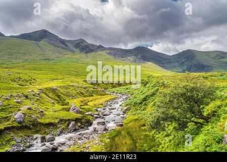 Winding mountain stream or river in Cronins Yard. Green fields in a valley with tall mountains covered in sunlight, Ring of Kerry, Ireland Stock Photo