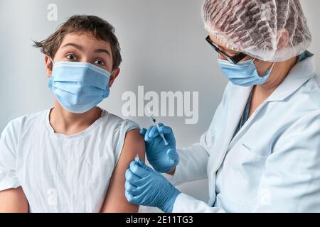 Anxious kid making face, scared of the syringe. Medic, doctor, nurse, health practitioner in white gown and face mask vaccinates teenage boy. Stock Photo