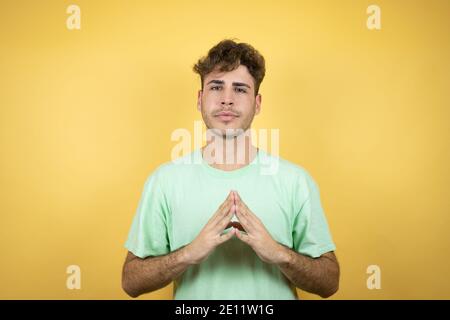 Handsome man wearing a green casual t-shirt over yellow background with Hands together and fingers crossed smiling relaxed and cheerful. Success and o Stock Photo