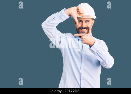 Young handsome man wearing architect hardhat smiling making frame with hands and fingers with happy face. creativity and photography concept. Stock Photo
