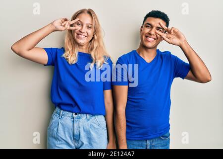 Young interracial couple wearing casual clothes doing peace symbol with fingers over face, smiling cheerful showing victory Stock Photo