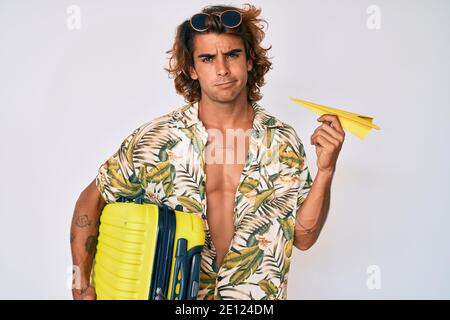 Young hispanic man holding suitcase and paper airplane going on summer vacation clueless and confused expression. doubt concept. Stock Photo