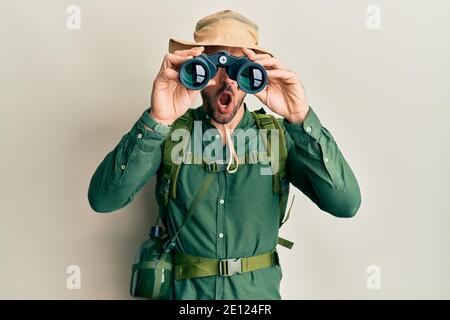Handsome man with beard wearing explorer hat looking through binoculars in shock face, looking skeptical and sarcastic, surprised with open mouth Stock Photo
