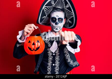 Young man wearing mexican day of the dead costume holding pumpkin pointing with finger to the camera and to you, confident gesture looking serious Stock Photo