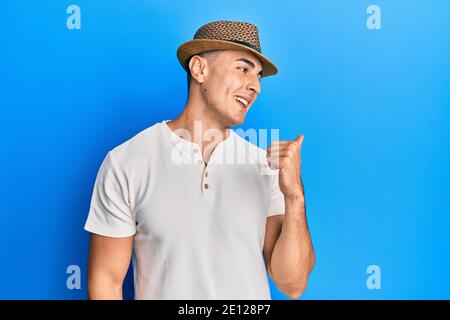 Hispanic young man wearing summer hat smiling with happy face looking and pointing to the side with thumb up. Stock Photo