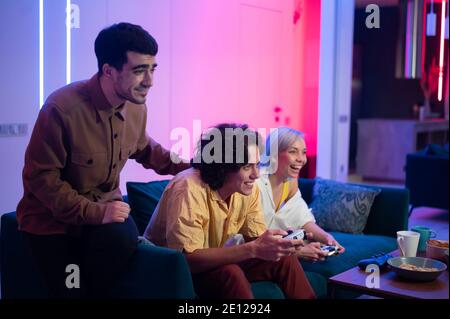 Company of the friends enjoy relaxing on couch playing videogames and having fun in modern flat lit with neon color. Stock Photo