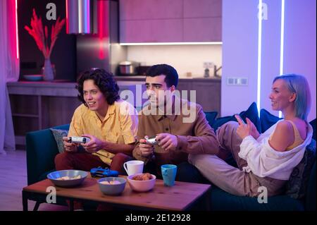 Friend of a young couple comes to their home to play games on gaming console on pleasant evening. Happy people playing video games at home. Home party Stock Photo
