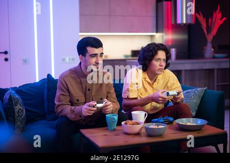Happy male gamers playing video games at home. Guy with manicure holding gaming console joystick. Stock Photo