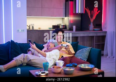 Man plays on gaming console using joystick while woman using smart phone with gaming app. Millenial couple lay on a couch at home and play video games Stock Photo
