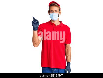 Handsome blond man with beard wearing delivery uniform and medical mask smiling happy pointing with hand and finger to the side Stock Photo