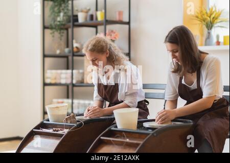 Two women make pottery on a pottery wheels, shaping clay by their hands, close up. Pottery craft workshop at art studio. Stock Photo