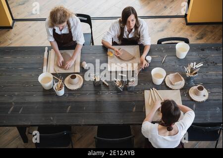 In the clay workshop. Female potters talking and smiling during the working process in the clay workshop. Female potter masters rolling up the clay on Stock Photo