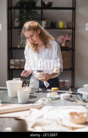 A young european beautiful female potter in a creative process of making ceramics at table in workshop. Hair fluttering in the wind at sunny day at Stock Photo