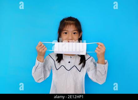 A cute Asian girl putting on a white cloth mask, covering her mouth and nose before going out to protect against Covid-19, with plain light blue backg Stock Photo