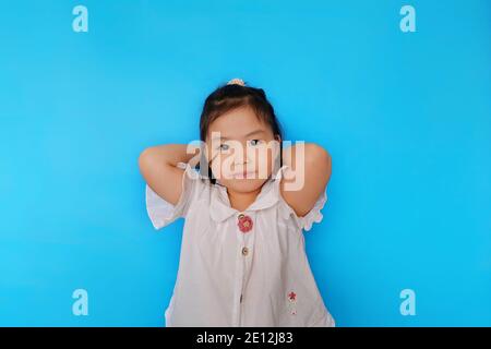 A cute young confident Asian girl is standing with her hands behind her head, smiling. Plain light blue background. Stock Photo