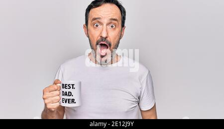 Middle age handsome man drinking cup of coffee with best dad ever message scared and amazed with open mouth for surprise, disbelief face Stock Photo