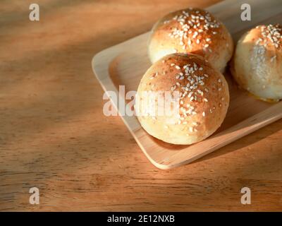 Fresh homemade burger buns with white sesame seed on small wooden tray on wood table with copy space. Three tasty sweet sandwich round buns just baked Stock Photo