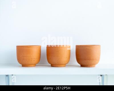 Three empty small modern terracotta pots on white wood shelf isolated on white wall background with copy space. Stock Photo