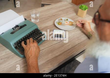Man enjoying eating fresh colorful asian sushi using chopsticks. This man writing with the old typewriter in the office. Stock Photo