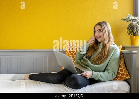 Young blonde girl using laptop computer on her bed in fornt of a yellow wall Stock Photo