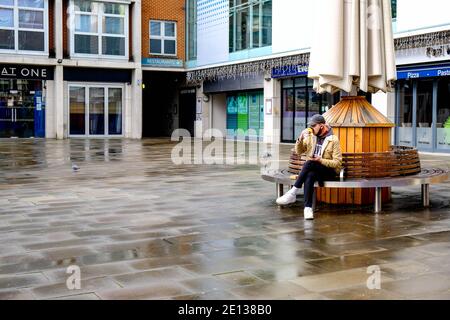 Epsom, London UK, January 03 2021, Lone Young Man Sitting Alone In An empty town Square Drinking Takeaway Coffee On A Wet Winters Day Stock Photo