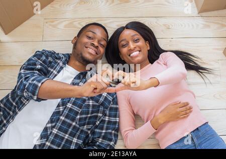 Loving black couple making heart with their hands, lying on floor surrounded by boxes on relocation day, top view Stock Photo