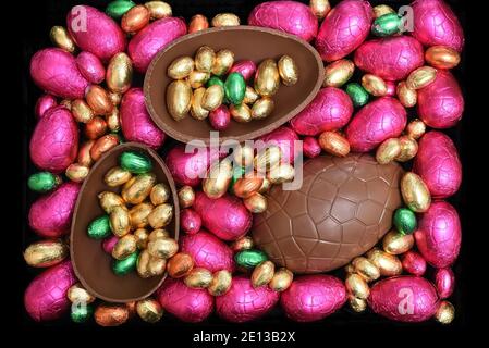 Pile or group of multi colored and different sizes of colourful foil wrapped chocolate easter eggs in pink, red, gold and lime green with two halves o Stock Photo