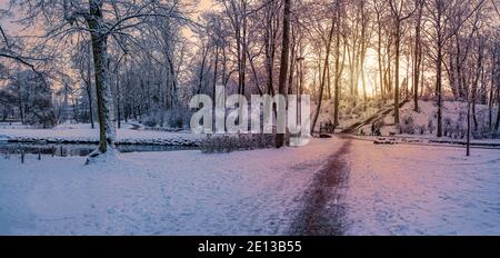 Panoramic view of sunset over  covered in snow park in winter. Small pond and stairs in snowy public park. Stock Photo