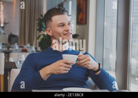Attractive man smiling, looking in the window, relaxing at the cafe holding cup of delicious coffee. Happy man having breakfast at the restaurant look Stock Photo