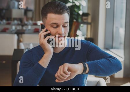 Handsome businessman talking on his phone, looking at the watch while waiting for a business meeting at the restaurant. Attractive man calling someone Stock Photo