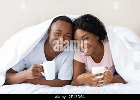 Happy black couple laying under blanket, drinking tea Stock Photo