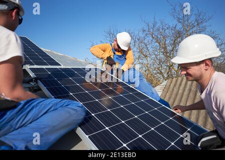 Male engineers installing solar photovoltaic panel system using screwdriver. Electricians mounting blue solar module on roof of modern house. Alternative energy ecological concept. Stock Photo