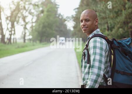 Attractive young African man hitchhiking on countryside road, smiling over his shoulder joyfully. Handsome cheerful man enjoying backpacking, walking Stock Photo