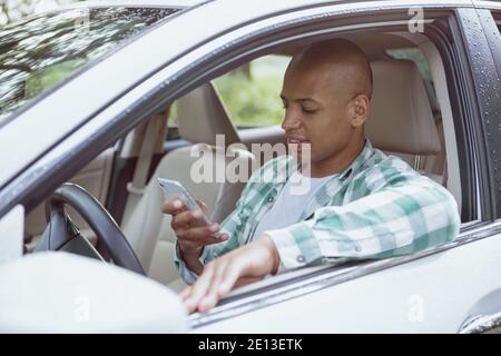 Young African handsome man using his smart phone, sitting in a car on countryside road, copy space. Attractive male driver using online map, travellin Stock Photo
