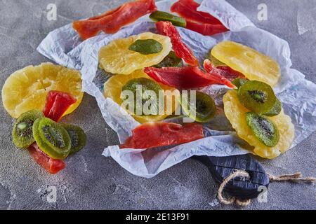 Dried fruits: Yellow candied pineapple rings, red papaya and green on white wrapping paper on gray concrete Stock Photo