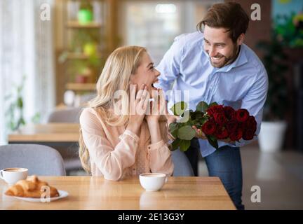 Celebrating special occasion. Romantic young guy presenting bouquet of red roses to his excited girlfriend at cafe Stock Photo